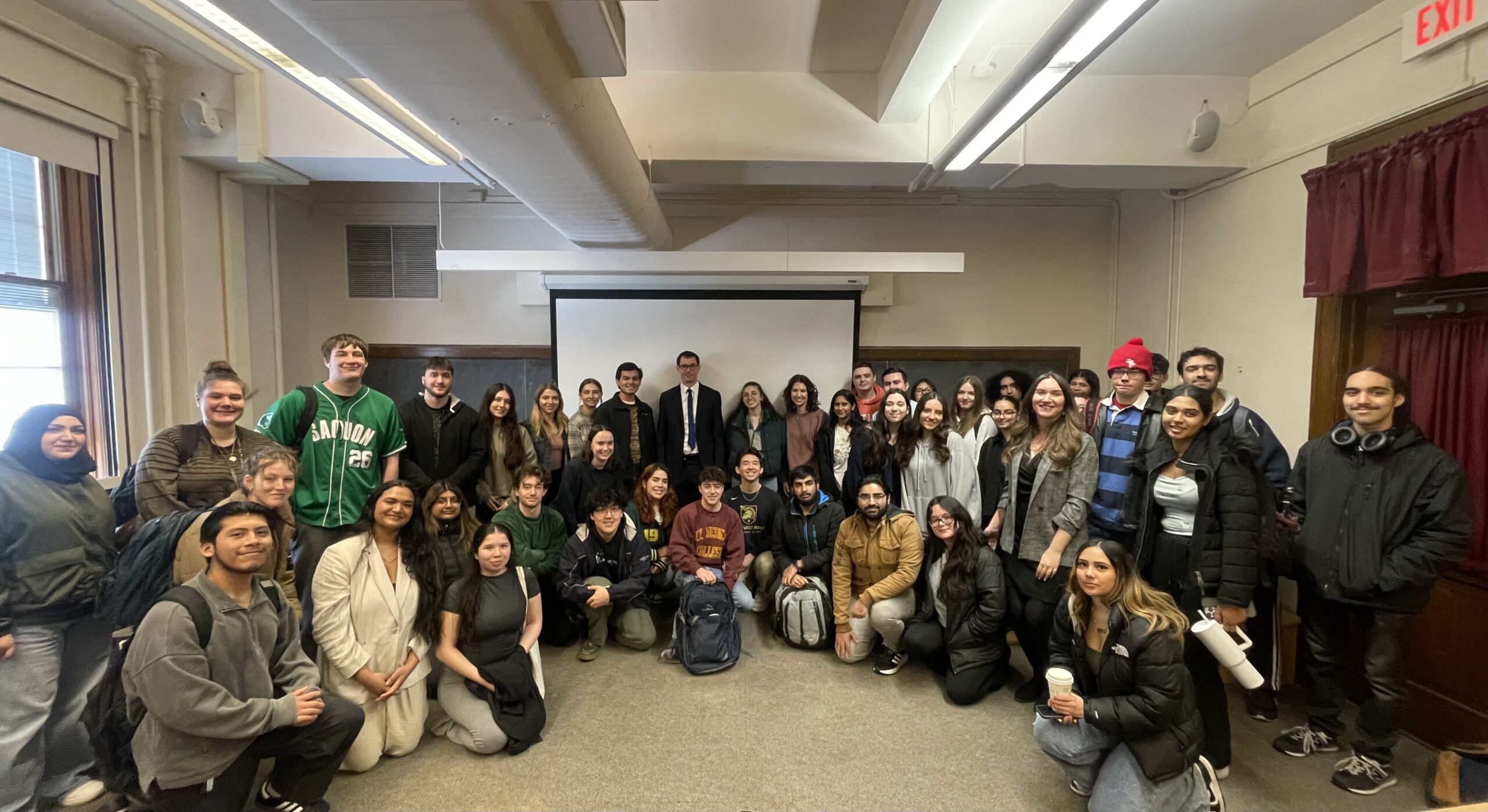 A large group of students and two instructors pose for a group photo in a classroom, smiling in front of a projection screen.